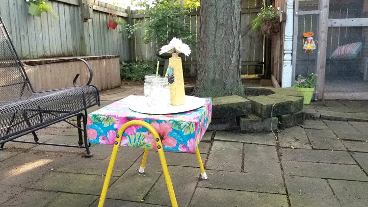 Colorful ottoman in the patio with a tray, water and an upcycled vase with daisies.