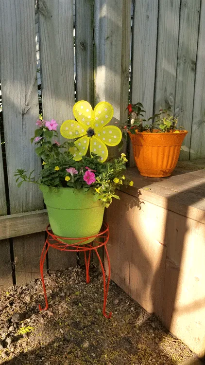 Green and orange painted plastic flower pots with a variety of flowers and a large yellow fabric flower that spins in the wind.