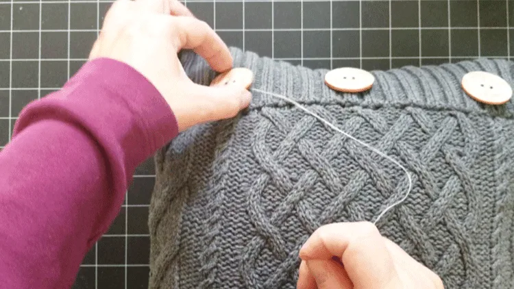 Hand stitching a wood button onto a gray sweater pillow