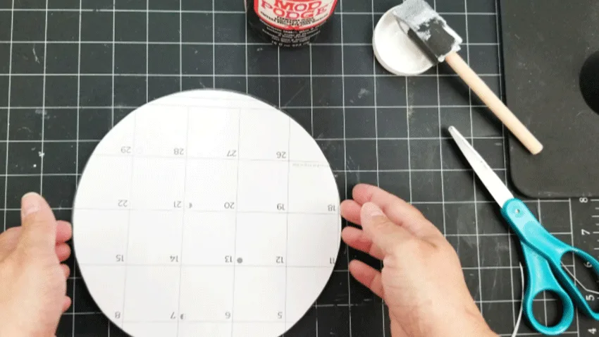Placing the cut calendar page onto the Mod Podge on the underside of the glass cutting board.